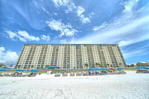 A large hotel with multiple floors and balconies, situated on a sandy beach with beach umbrellas and lounge chairs.