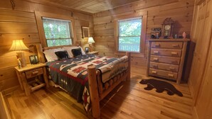 Un dormitorio de cabaña de madera con una cama, dos mesitas de noche, un aparador y una ventana con vistas a árboles.