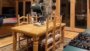 Interior de madera con chimenea de piedra, mesa y sillas de madera, y una zona de cocina con estufa.
