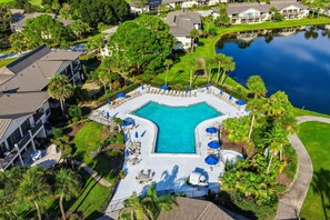 An aerial view of a resort with a central swimming pool surrounded by lounge chairs and umbrellas, set against a backdrop of residential buildings and a body of water.
