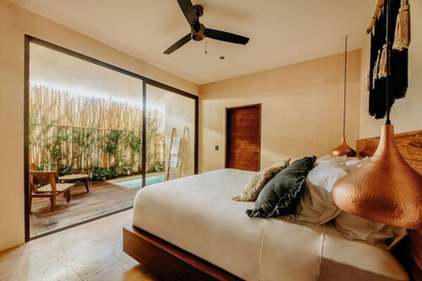 A bedroom with a bed, a ceiling fan, and a view of a bamboo fence outside. - Majaro Apartments (Tulum)