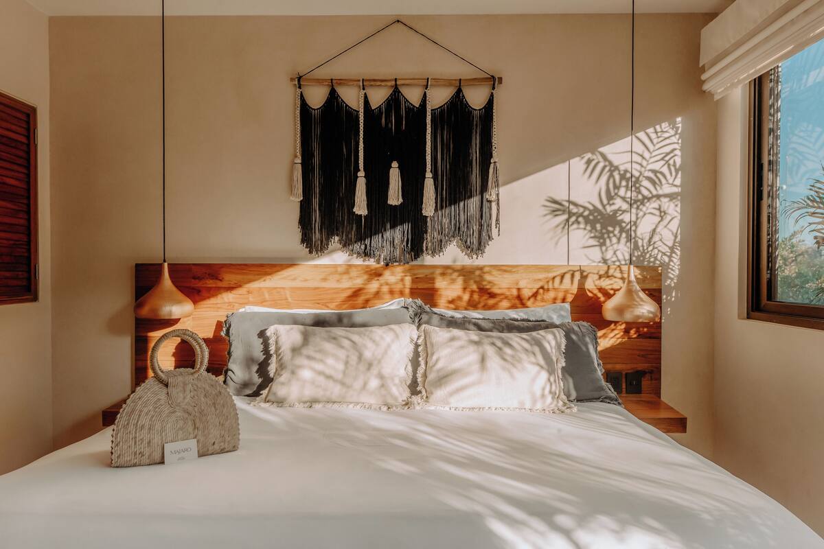 Un dormitorio con cama, un tapiz de macramé, cabecera de madera y lámpara colgante.