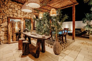 An outdoor seating area with a stone wall, wooden furniture, and hanging wicker lamps. - Majaro Apartments (Tulum)