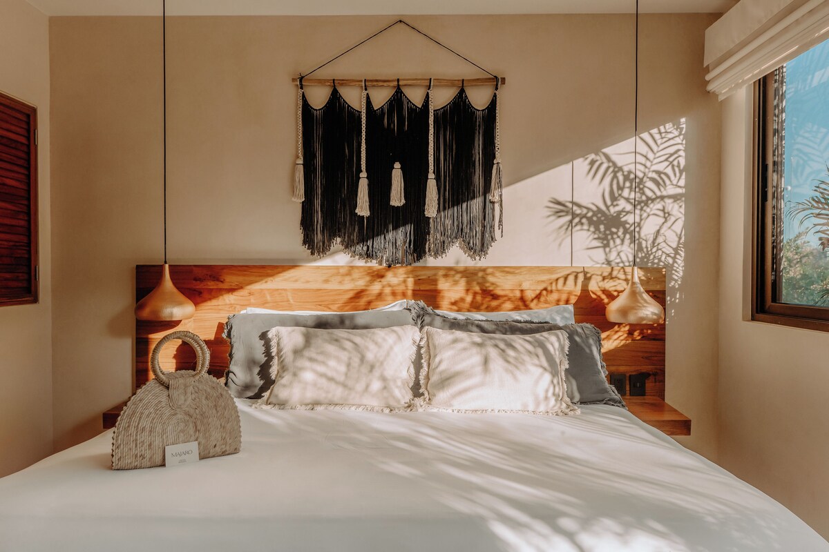 A neatly made bed with a headboard, a hanging macrame piece, and a small woven bag on the nightstand.