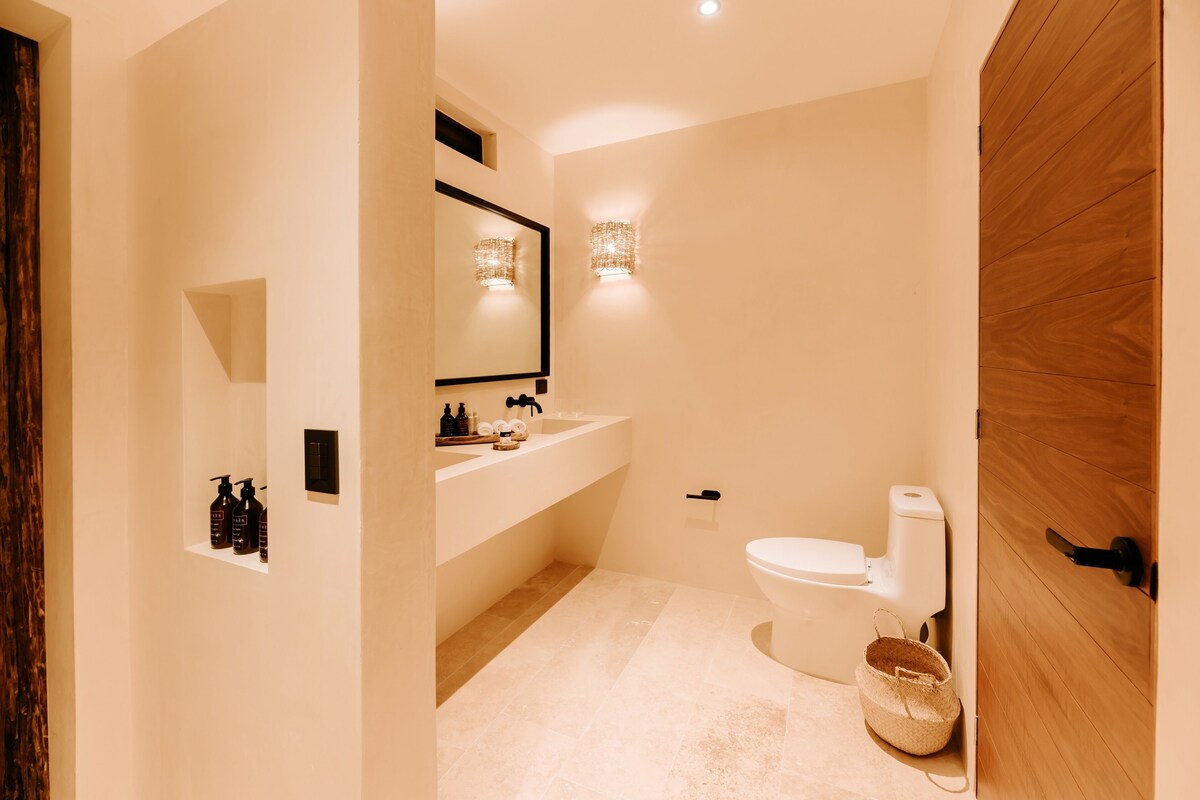A modern bathroom with a wooden door, a white toilet, a sink with a mirror above it, and a wall-mounted light fixture.