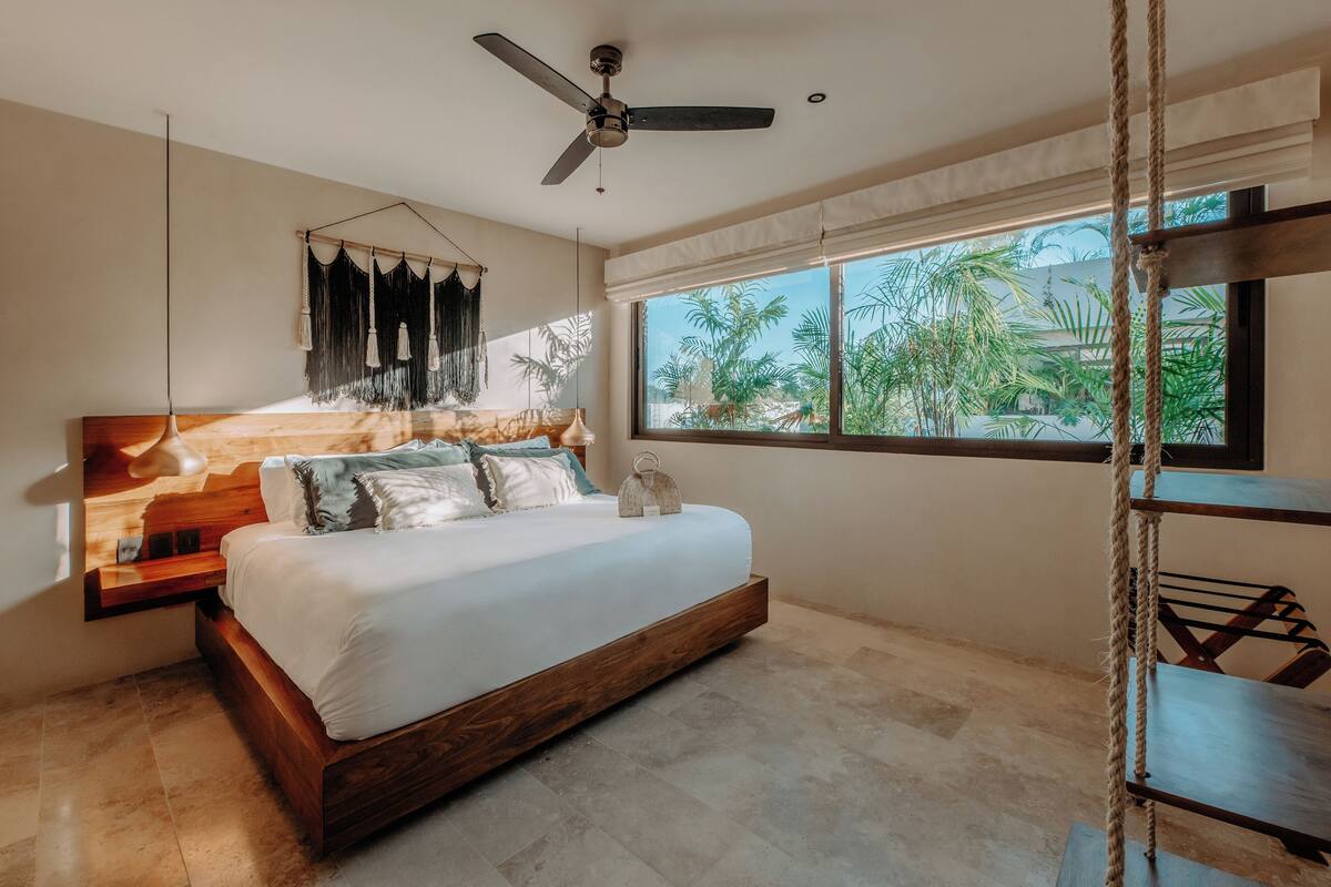 Un dormitorio moderno con cama de madera, ventilador de techo, ventana grande con vista a palmeras y decoración de macramé colgante.