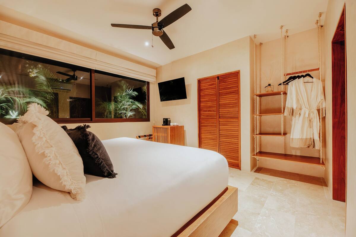 Un dormitorio con cama, ventilador de techo, televisor, ventana con vista a palmeras y un armario de madera.