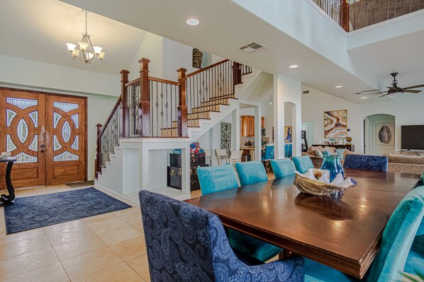 A spacious living room with a large wooden dining table, blue upholstered chairs, a staircase with wooden railings, and a chandelier.