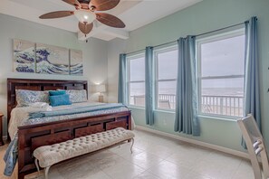 A bedroom with a large bed, a bench, a ceiling fan, and a view of the ocean through a window.