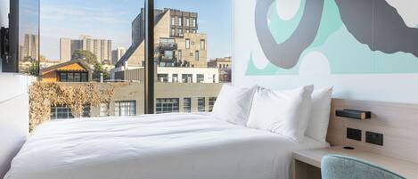 A modern hotel room with a large bed, a view of the cityscape, and a wall-mounted artwork.