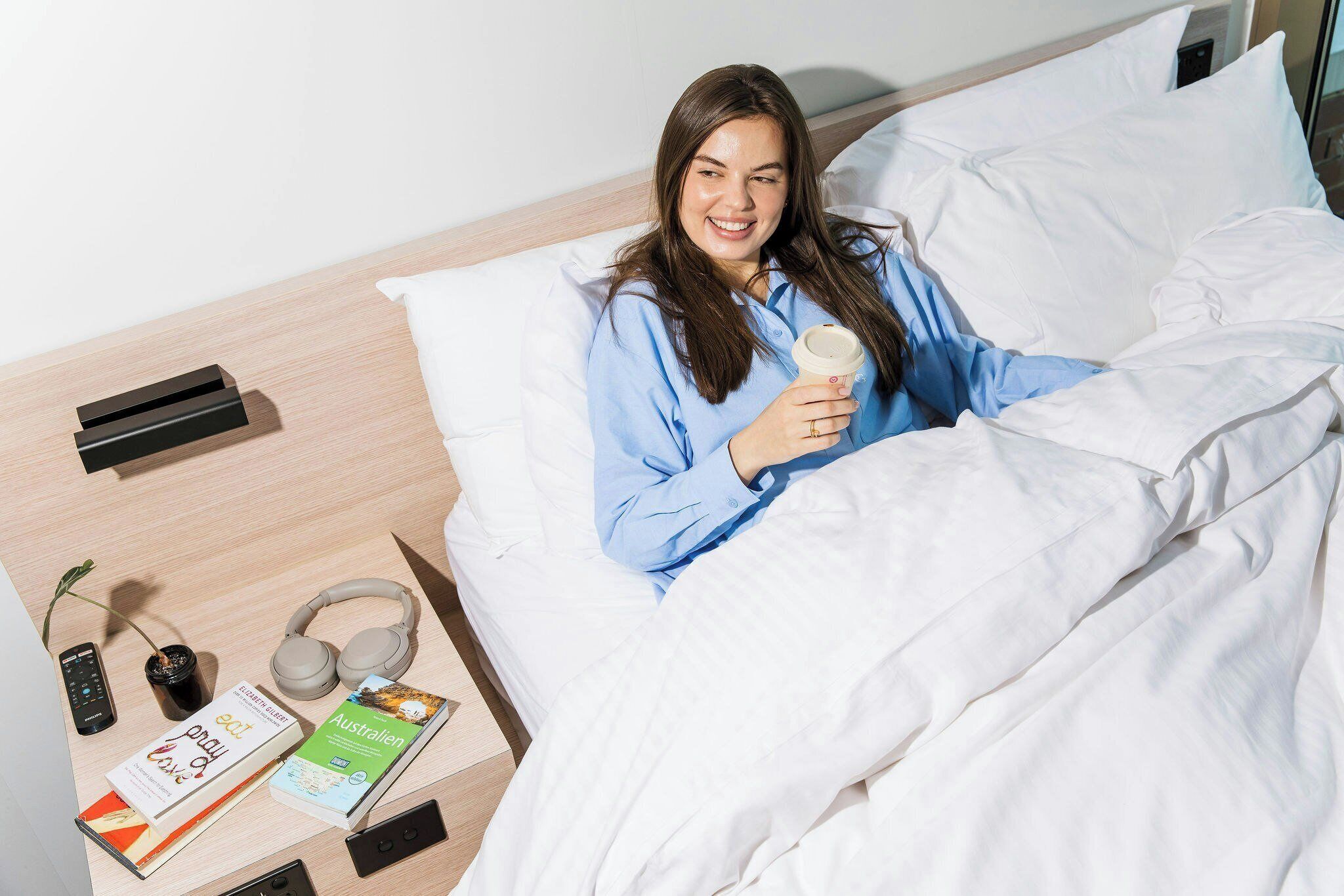 A person lying in bed with a book, headphones, and a remote control on a bedside table.