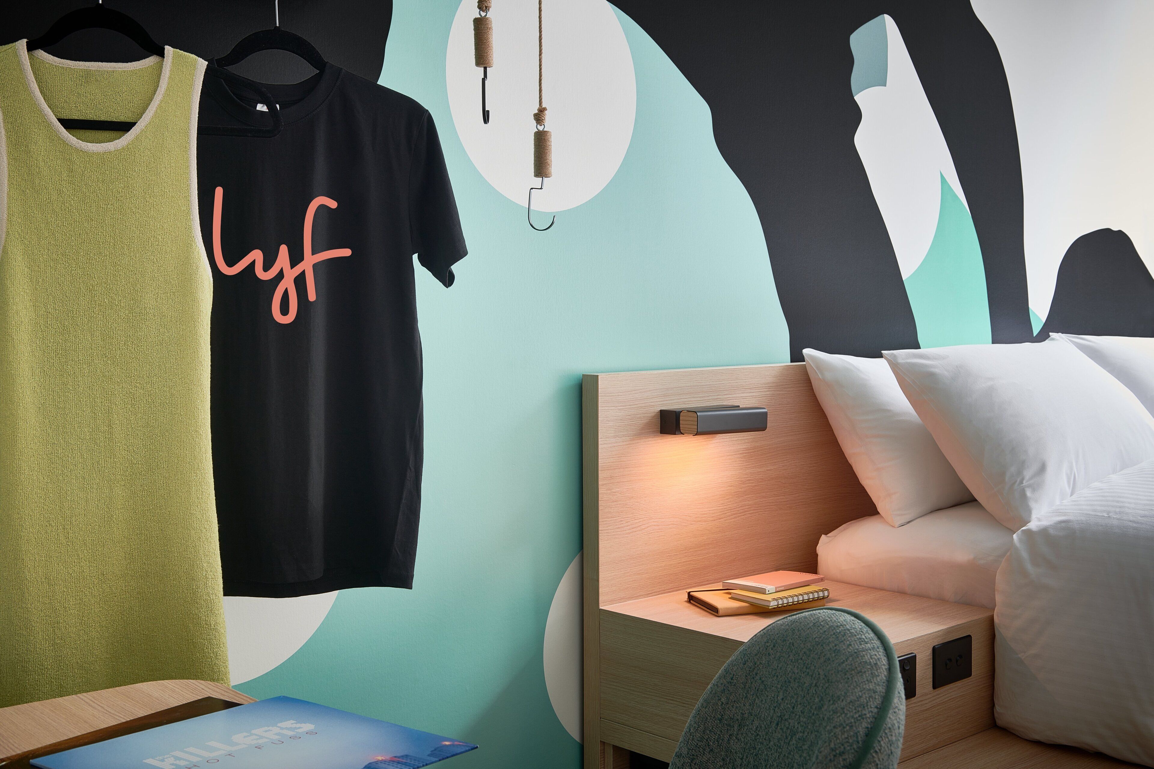 Un dormitorio con una cama, una mesita de noche con un libro, y dos camisas colgadas en un perchero.