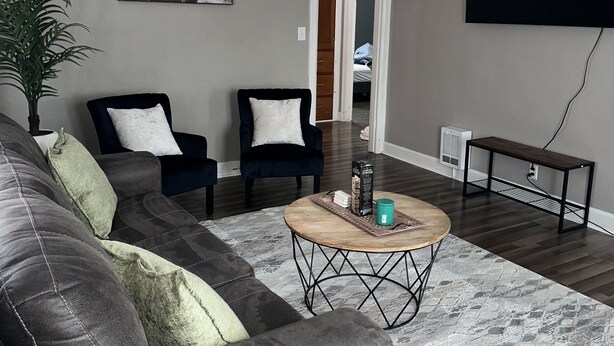 A living room with a dark gray sofa, two dark armchairs, a wooden coffee table, a ceiling fan, a mounted TV, and a framed picture on the wall.