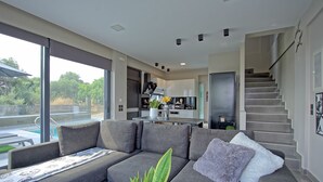 A modern living room with a grey sofa, a coffee table, and a potted plant. A staircase leads to a kitchen area with a refrigerator and a dining table.
