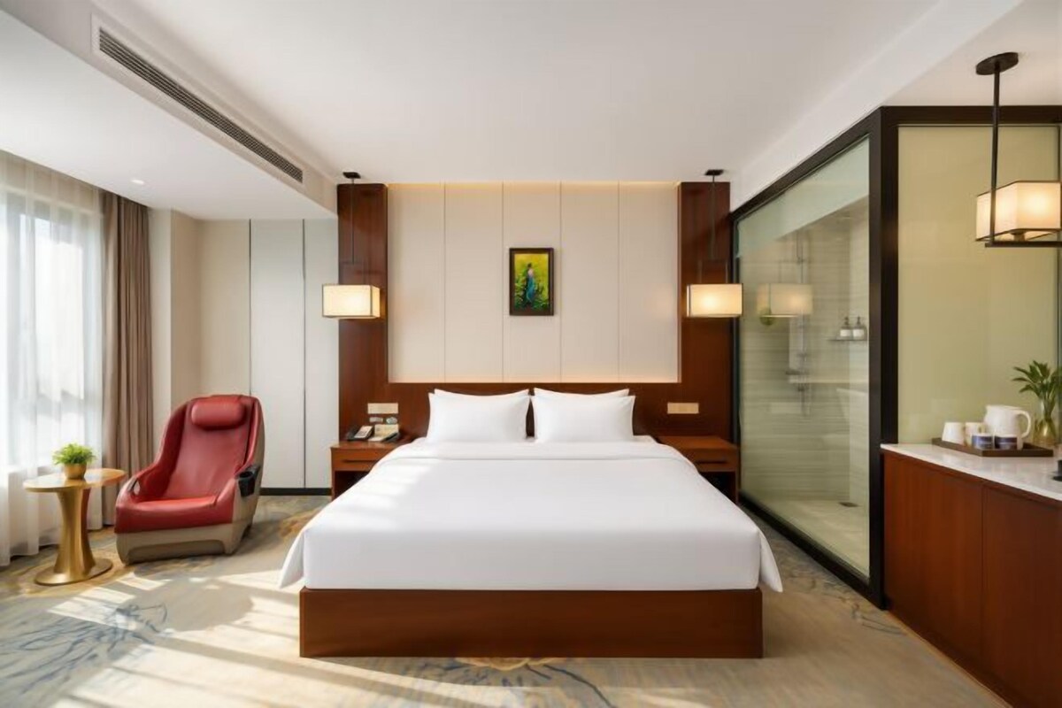 Ein Hotelzimmer mit einem großen Bett, einem roten Sessel, einem Schreibtisch mit Lampe und Blick auf die Stadt durch eine Glaswand.