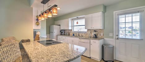 A kitchen with white cabinets, a granite countertop, and a built-in stove. There is a trash can and a window with blinds.