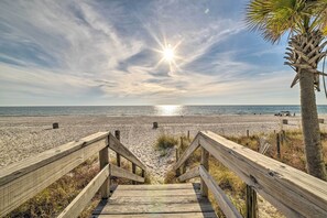 Een houten trap die naar een strand leidt, met een palmboom en de zon die in de lucht schijnt.
