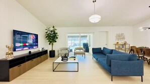 Ett modernt vardagsrum med en platt-TV, en stor krukväxt, en blå soffa och en matplats med stolar och ett bord.