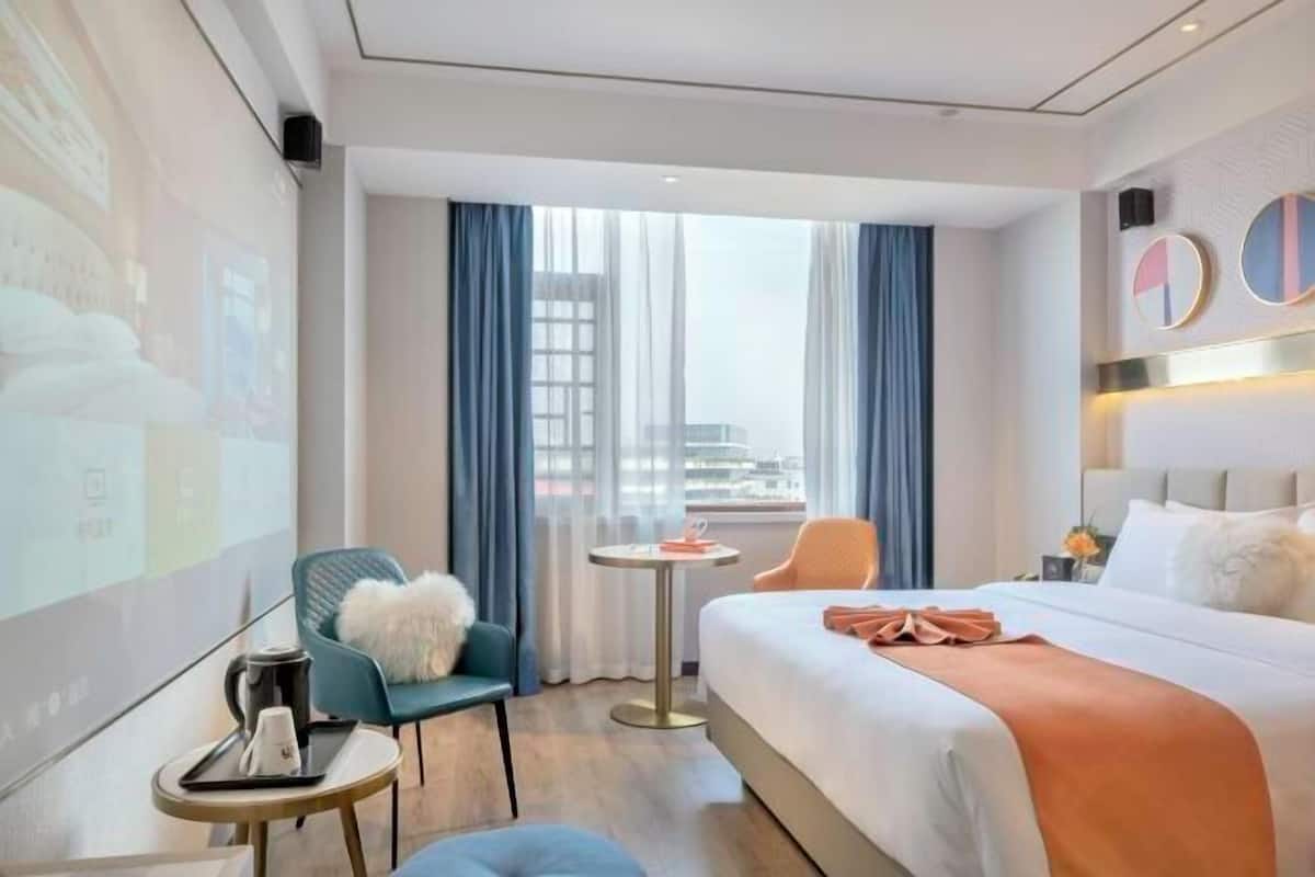 A modern hotel room with a large bed, a small table, a chair, and a view of the city through large windows.