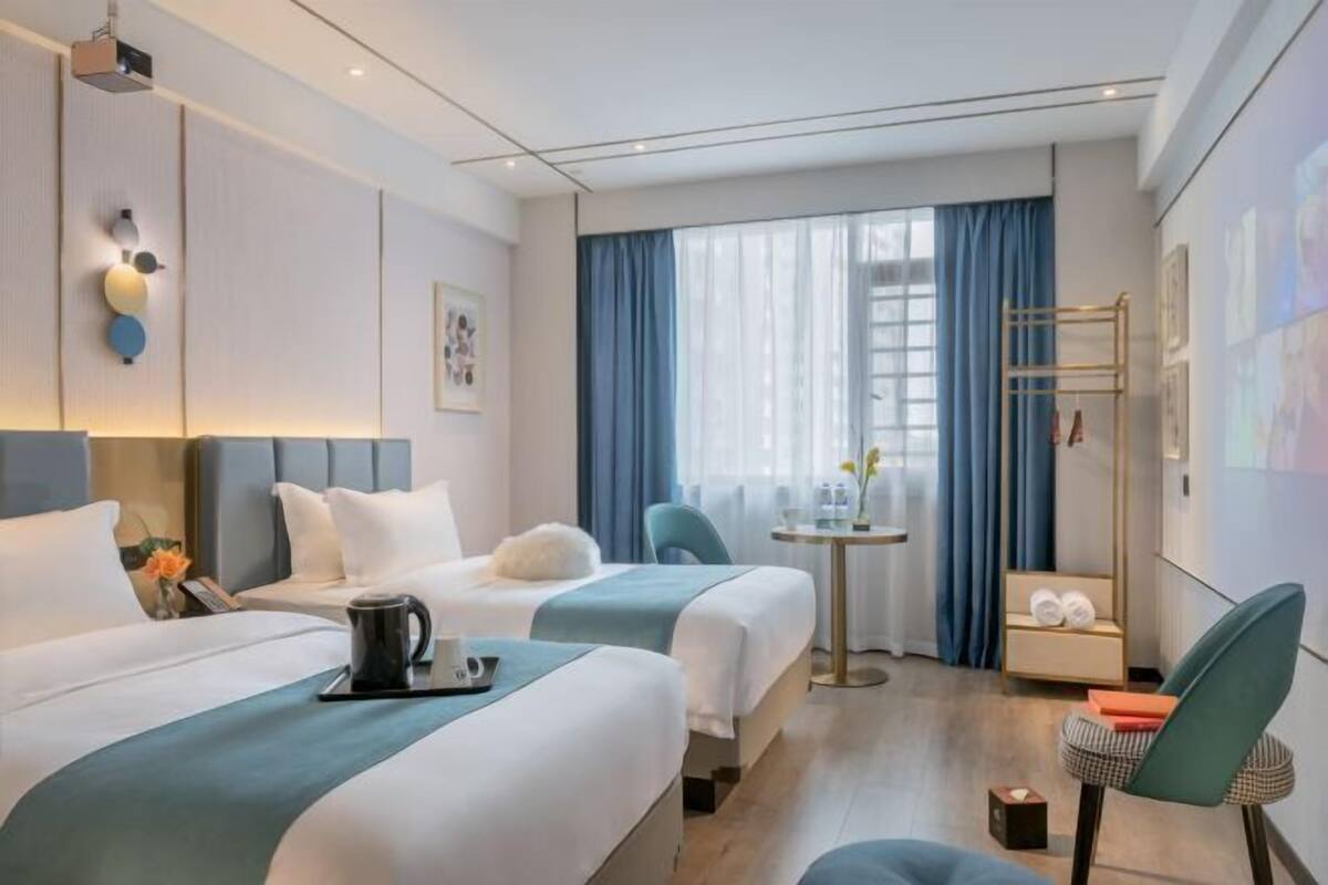 A hotel room with two beds, a small table, a chair, and a window with curtains.