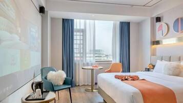 A modern hotel room with a large bed, a small table, a chair, and a view of the city through large windows.