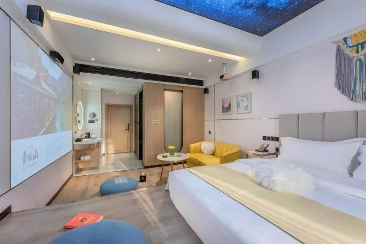A modern hotel room with a large bed, a sofa, a small table, and a ceiling projector displaying a scenic image.