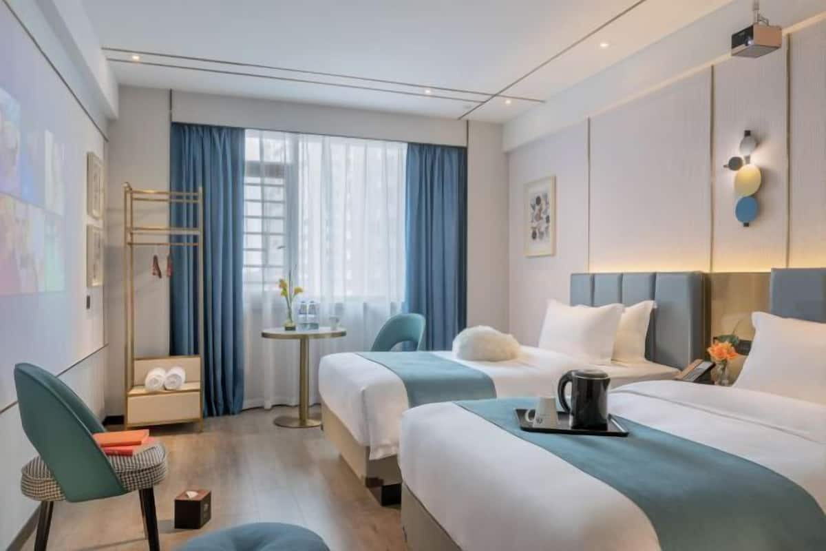 A hotel room with two beds, a small table with a kettle, a chair, and a window with curtains.
