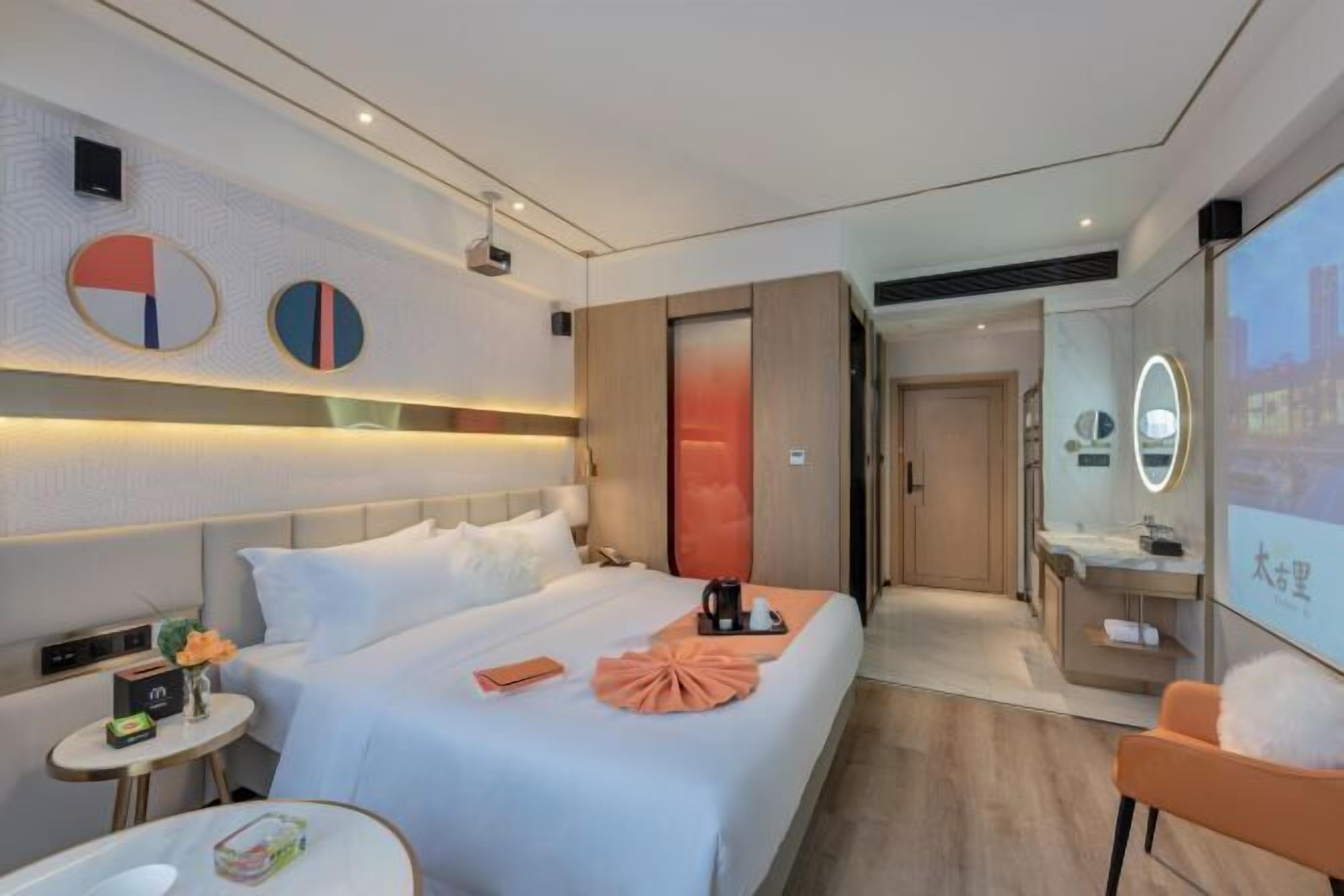 A modern hotel room with a neatly made bed, a small table with a vase of flowers, a mirror, and a view of the cityscape through the window.