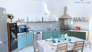 A kitchen with a built-in oven, a coffee machine, and a sink. There is a dining table with chairs and a tablecloth.