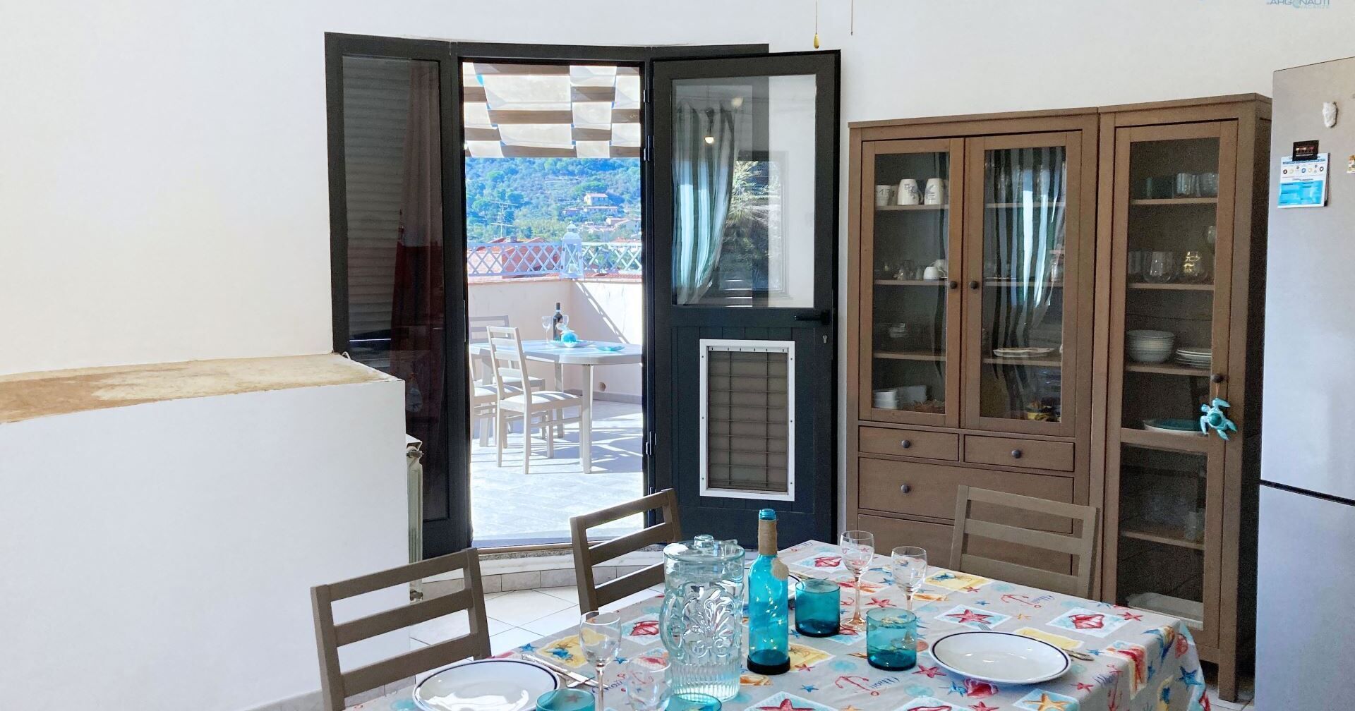 A dining area with a table set for two, a cabinet with glass doors, and a view of a balcony with outdoor furniture.