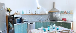 A kitchen with a built-in oven, a coffee machine, and a sink. There is a dining table with chairs and a tablecloth.