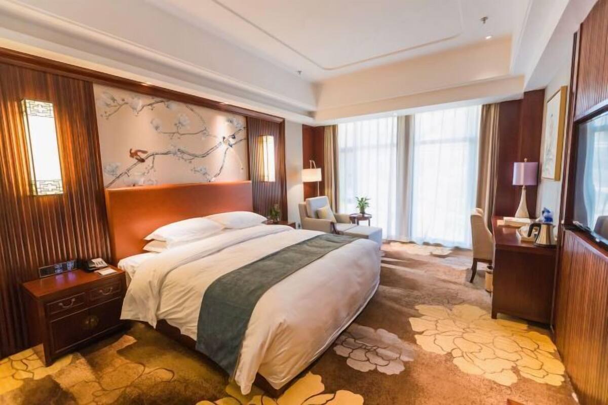 A hotel room with a large bed, a desk, a chair, a nightstand, a lamp, and a wall with a floral pattern.