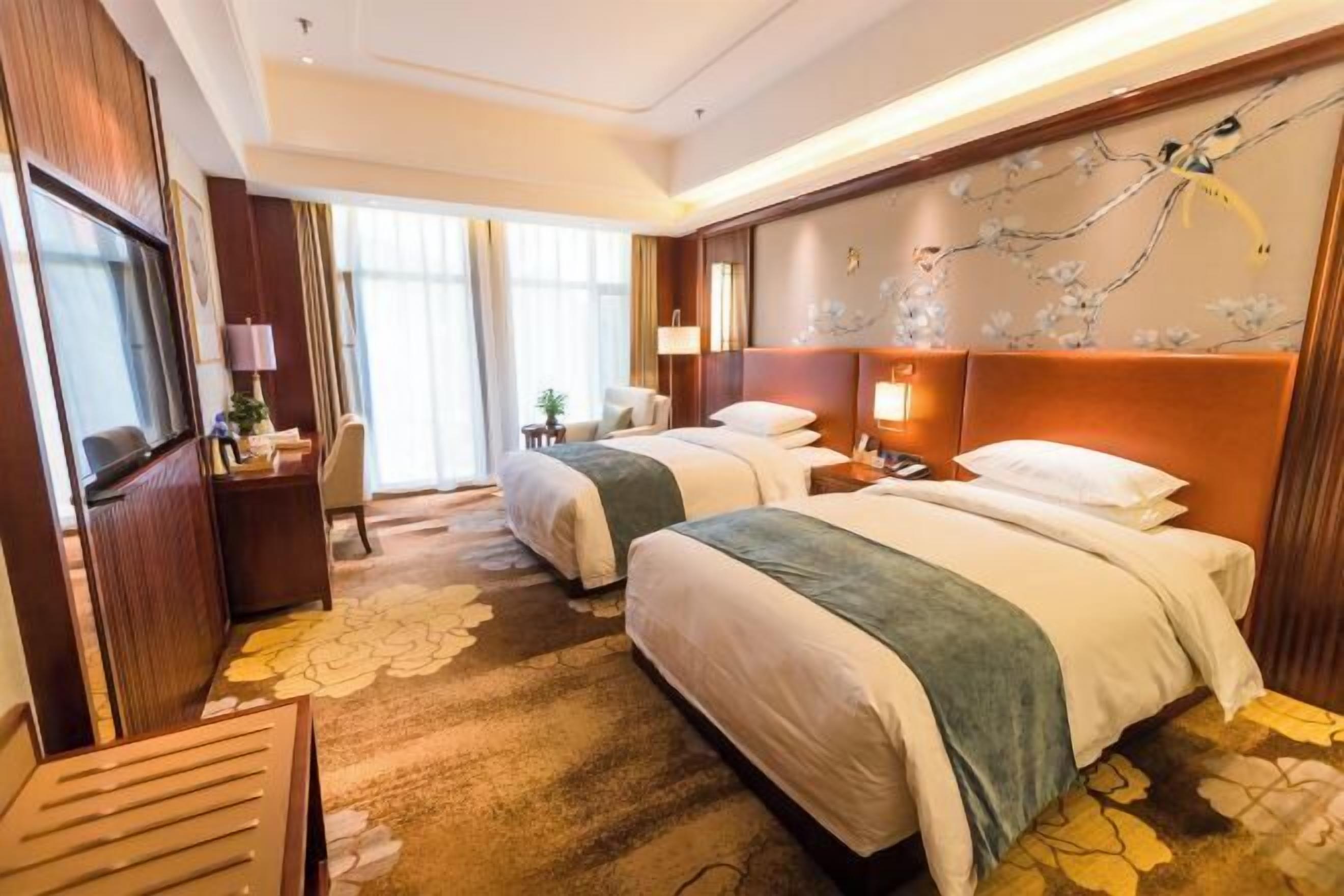 Quarto de hotel com duas camas, uma escrivaninha, uma cadeira, uma televisão e uma janela grande com cortinas.