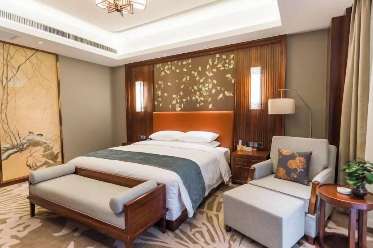 A hotel room with a large bed, a sitting area with a chair and a small table, a decorative wall panel, and a floor lamp.