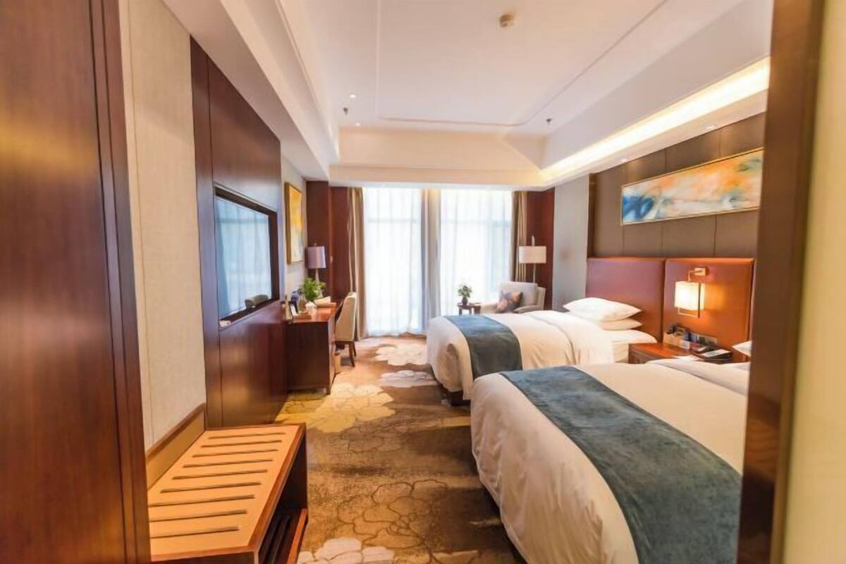A hotel room with two beds, a desk, a chair, a window, and a painting on the wall.
