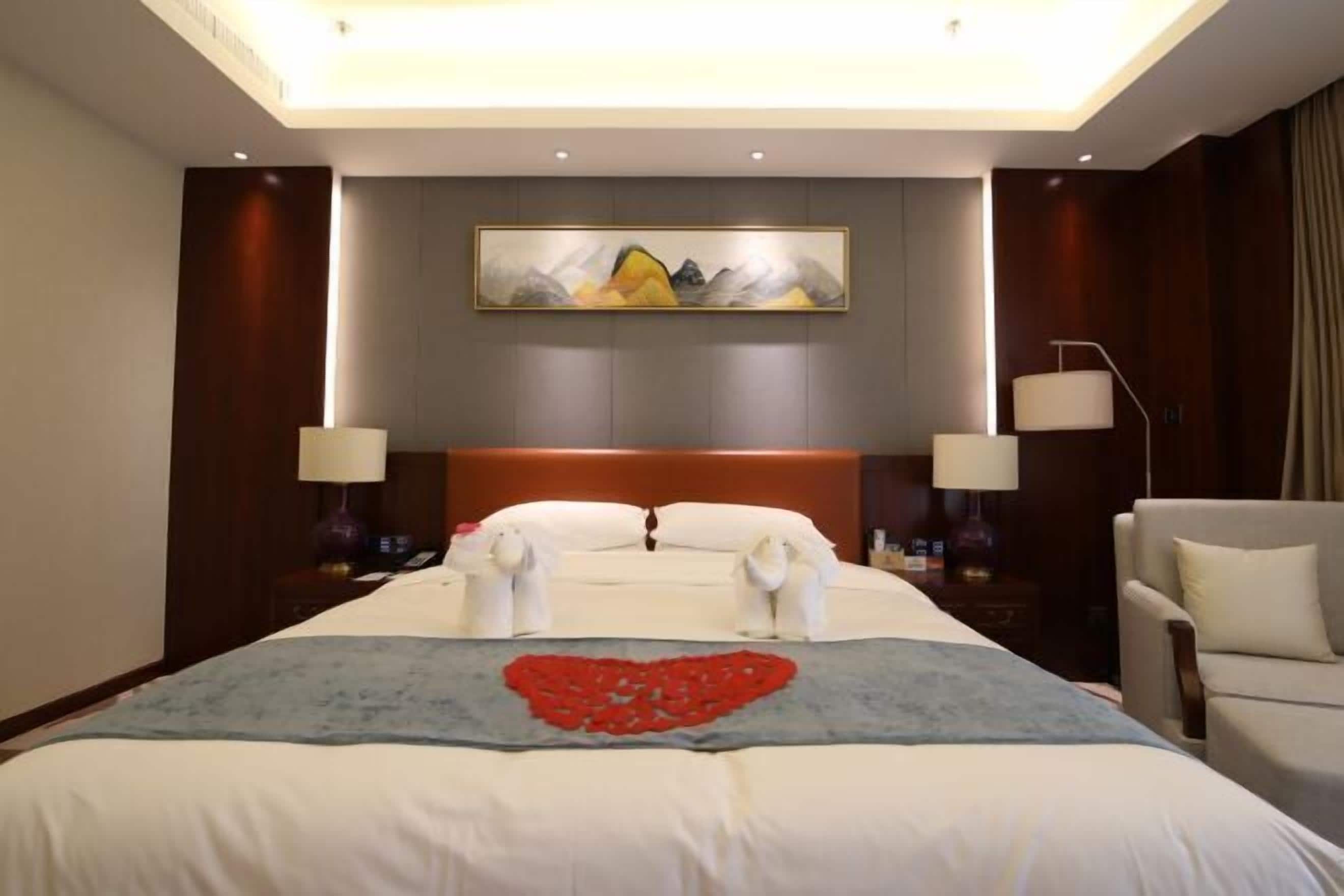 Habitación de hotel con una cama grande, un cuadro en la pared y una decoración con forma de corazón en la colcha.