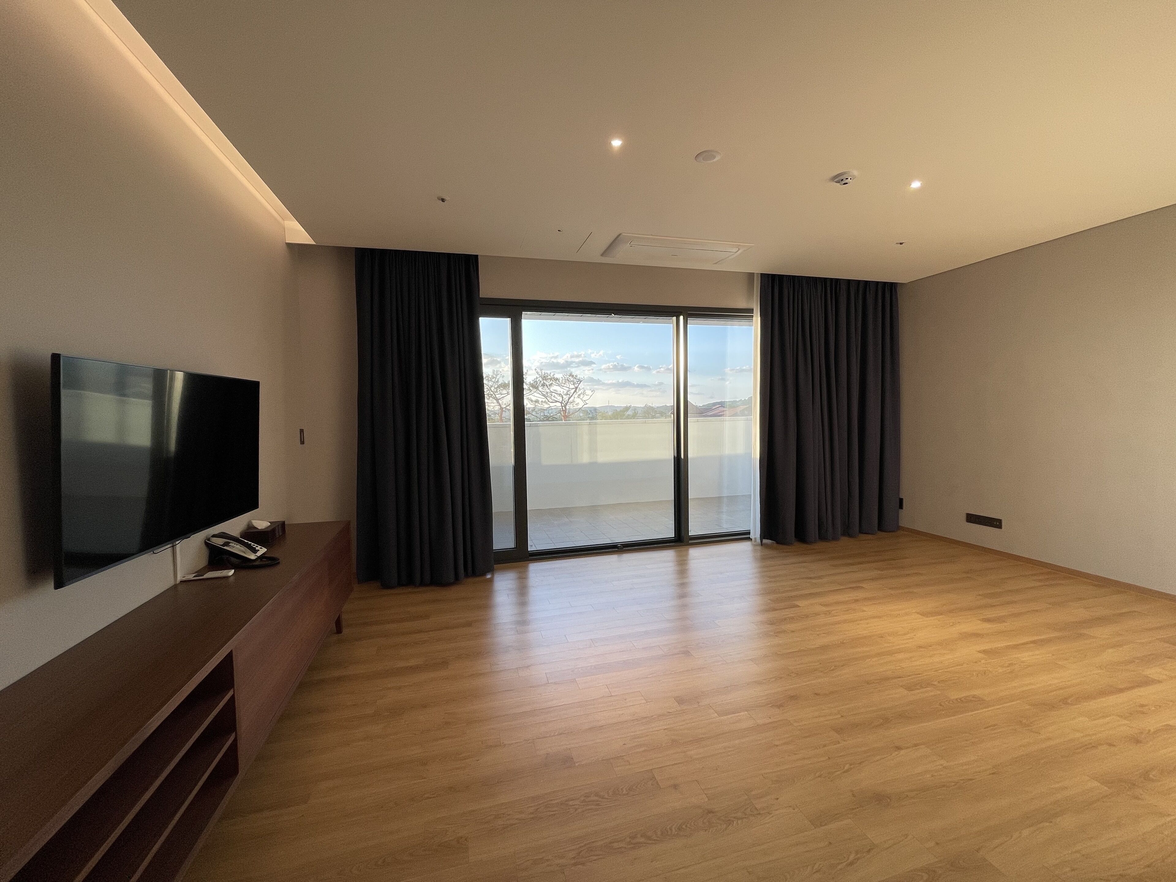 Una habitación amplia con televisión de pantalla plana, piso de madera y un gran ventanal corredero que da a un balcón.