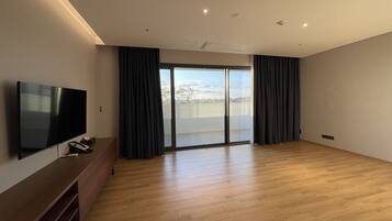 Una habitación amplia con televisión de pantalla plana, piso de madera y un gran ventanal corredero que da a un balcón.