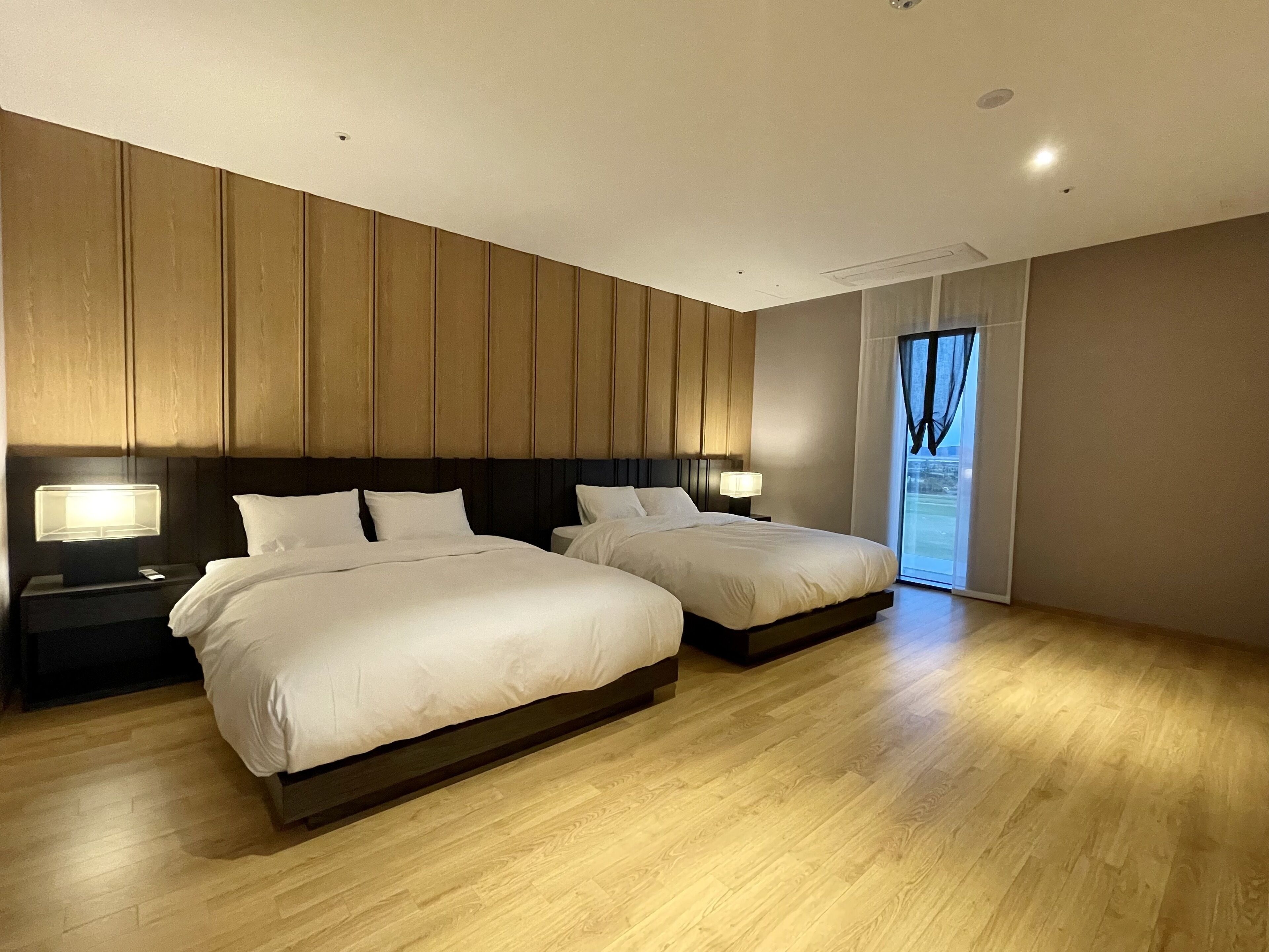 Quarto de hotel moderno com duas camas, painéis de madeira nas paredes e uma janela grande com cortinas.