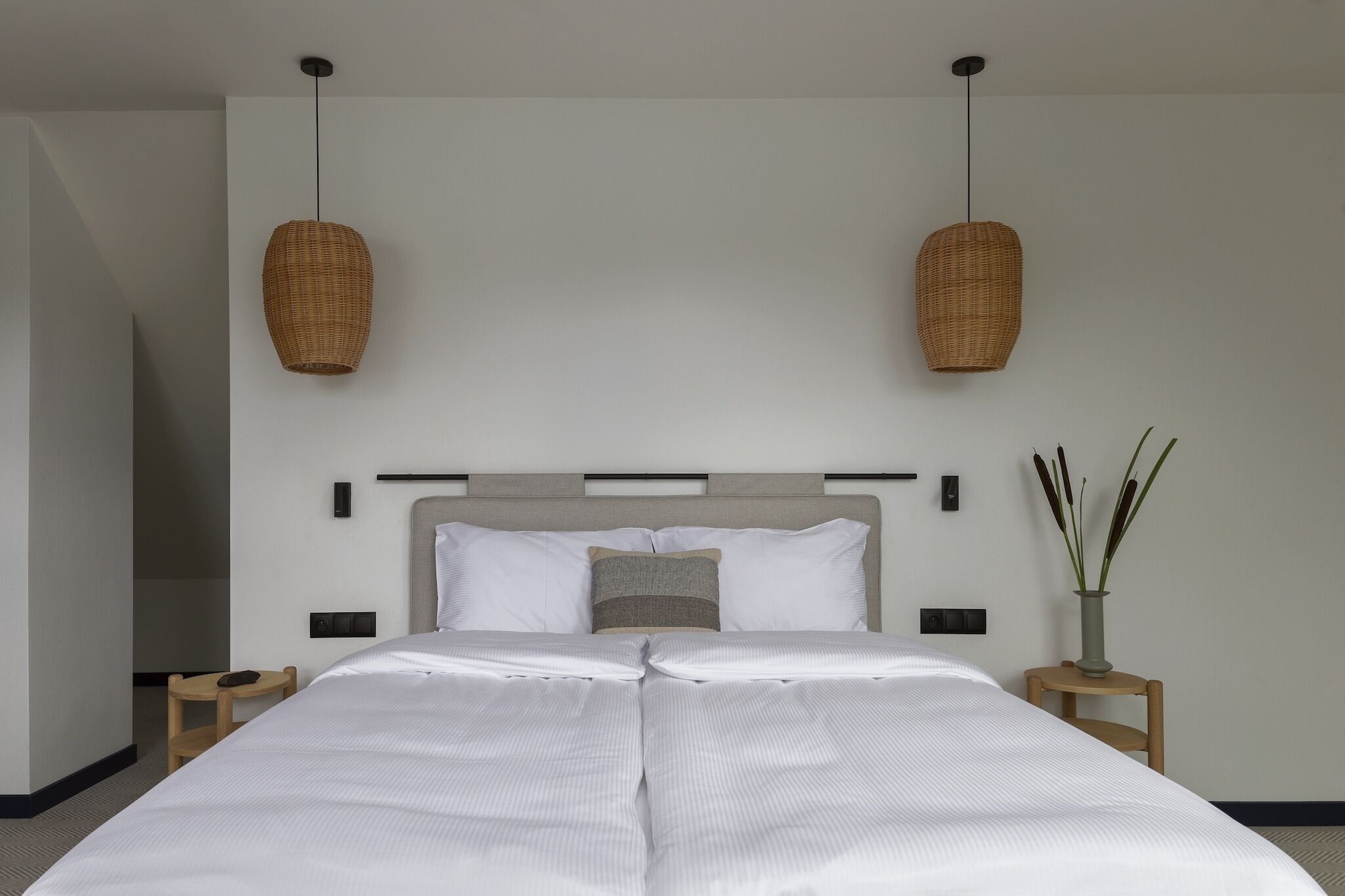 Quarto de hotel moderno com uma cama grande, duas mesas de cabeceira, um criado-mudo, um vaso com plantas e duas luminárias de vime suspensas.