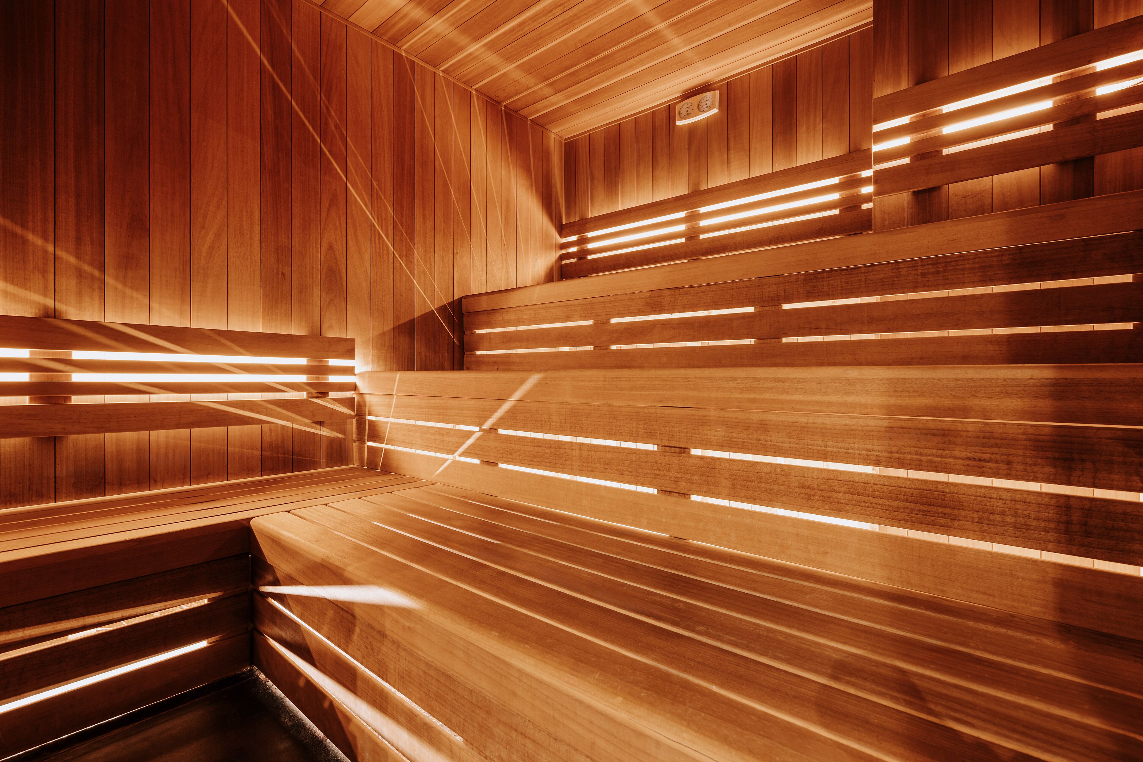 A sauna with wooden benches and slatted backrests, illuminated by linear lights.