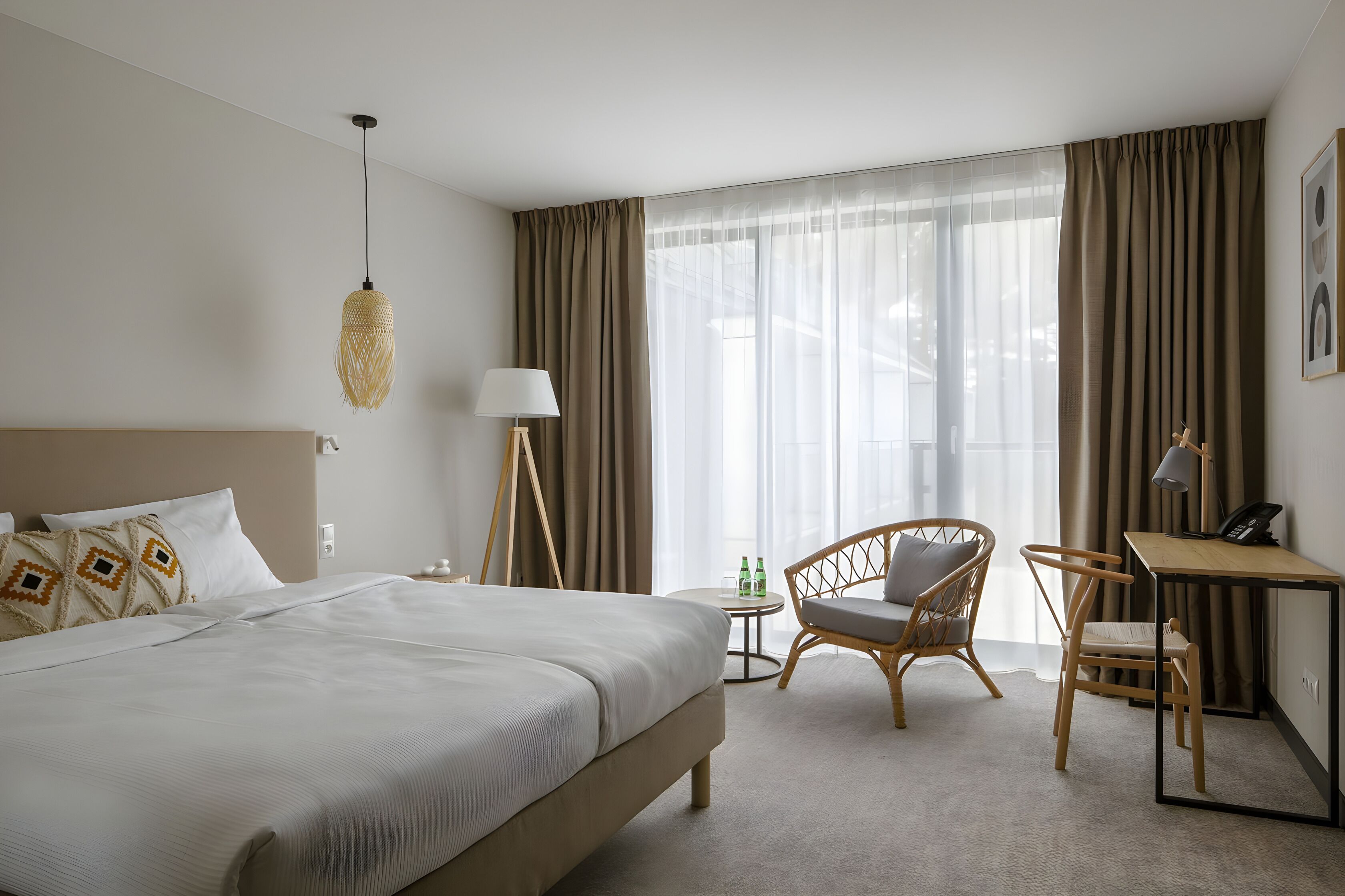 Quarto de hotel com uma cama grande, uma escrivaninha, duas cadeiras, um abajur e um lustre.