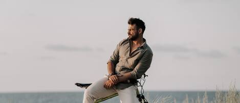 Een persoon zittend op een fiets in een zandrijk gebied met schaarse begroeiing en een heldere lucht.