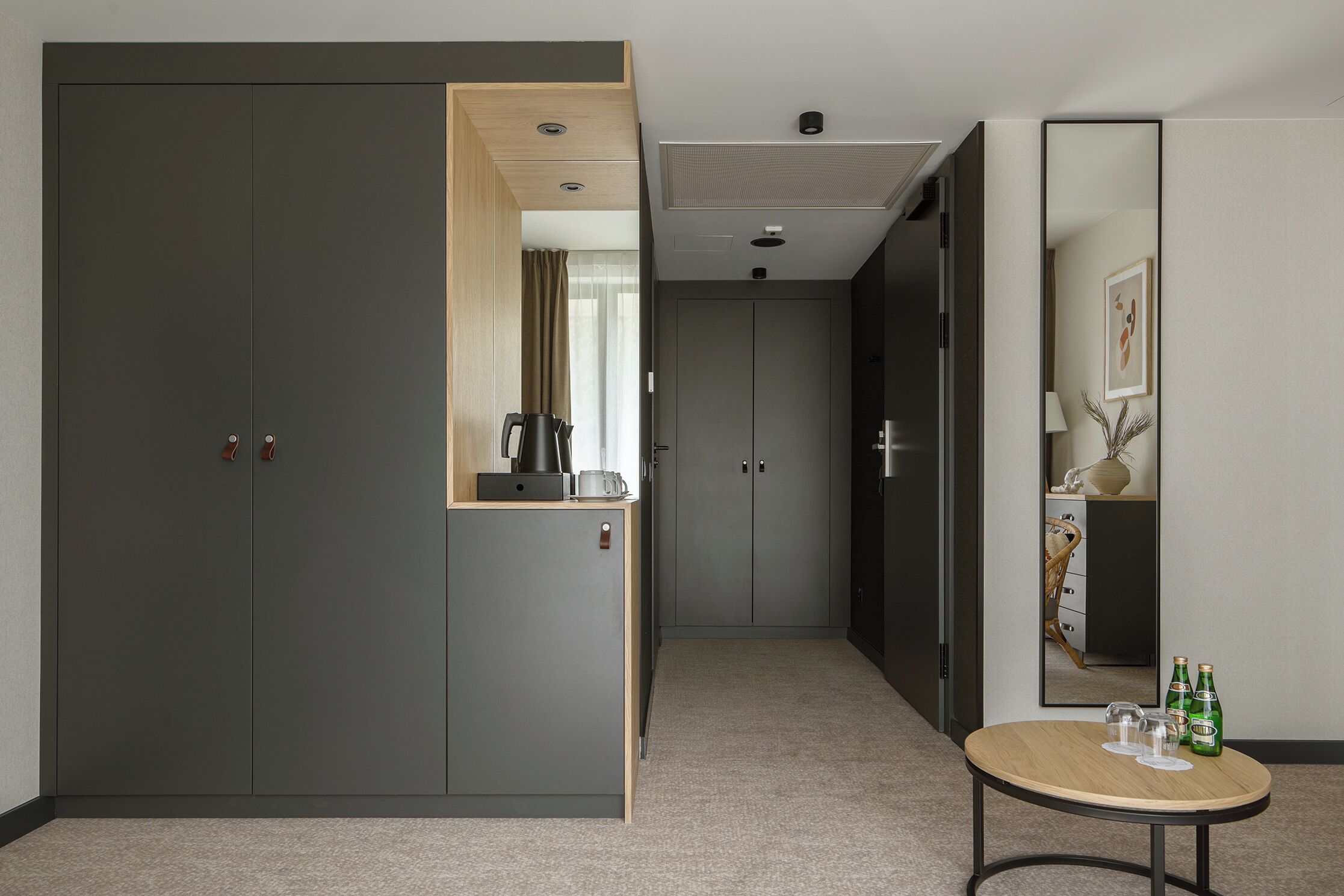 A modern hotel room with a large wardrobe, a small table with glasses and bottles, and a mirror reflecting the room.