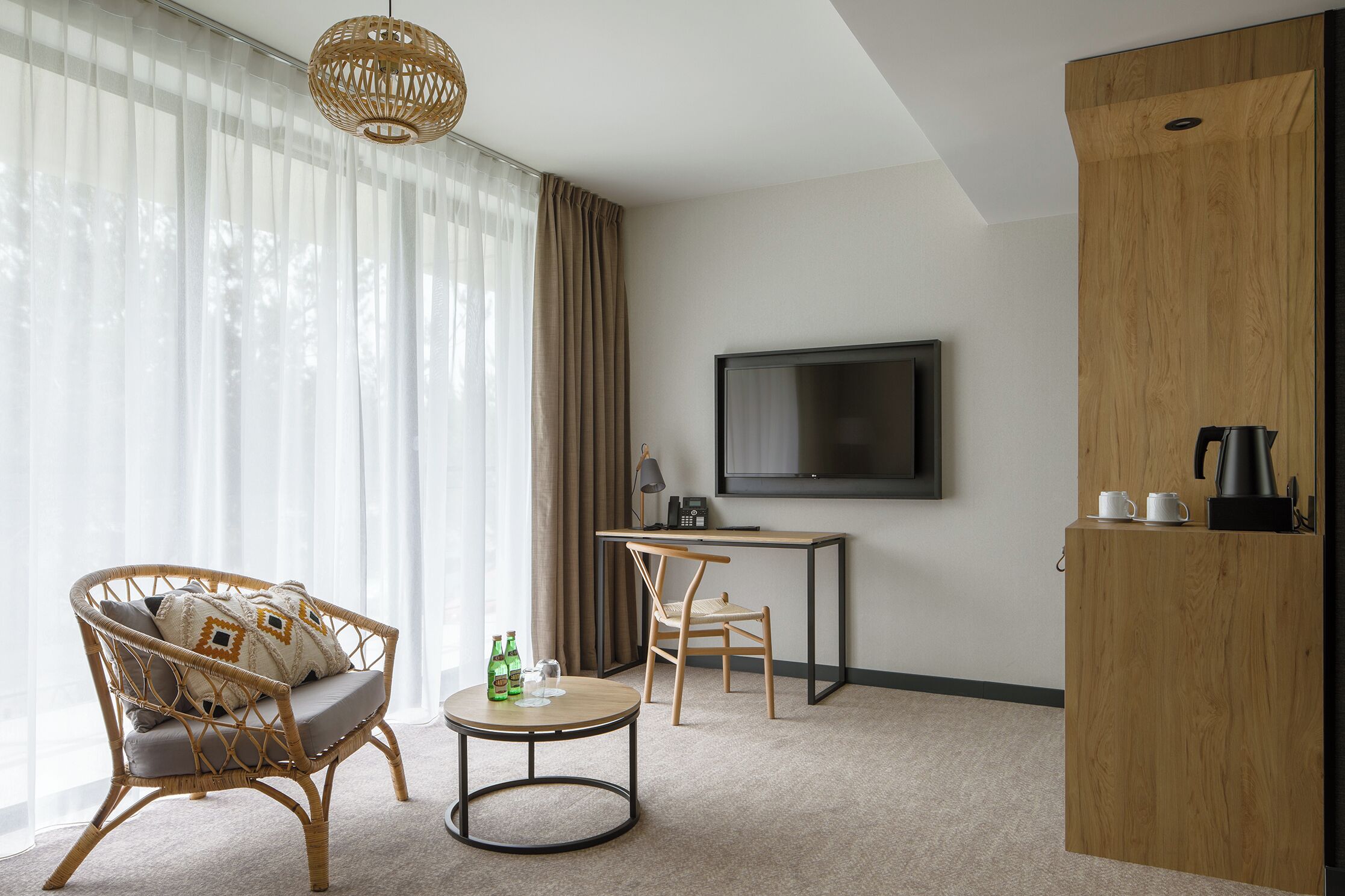 A modern hotel room with a large window, a wicker sofa, a small round table, a desk with a chair, a television, and a wooden cabinet.