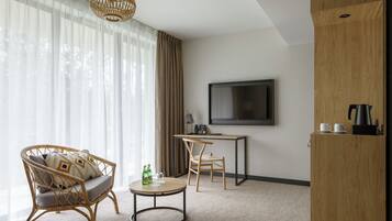 A modern hotel room with a large window, a wicker sofa, a small round table, a desk with a chair, a television, and a wooden cabinet.