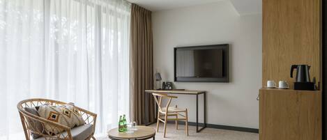 A modern hotel room with a large window, a wicker sofa, a small round table, a desk with a chair, a television, and a wooden cabinet.