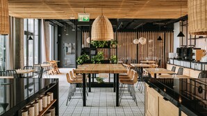 A modern restaurant interior with a central wooden table, black chairs, and a hanging wicker light fixture.