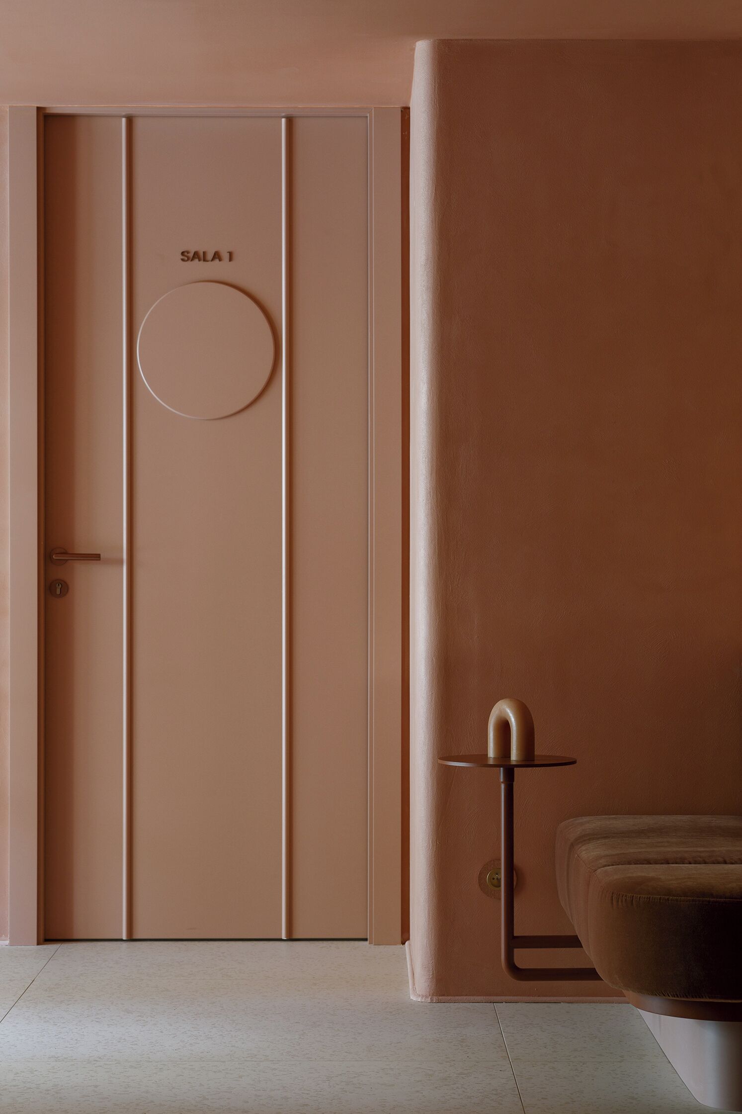 A room with a closed door labeled "SALA 1," a small table with a lamp, and a cushioned seating area.