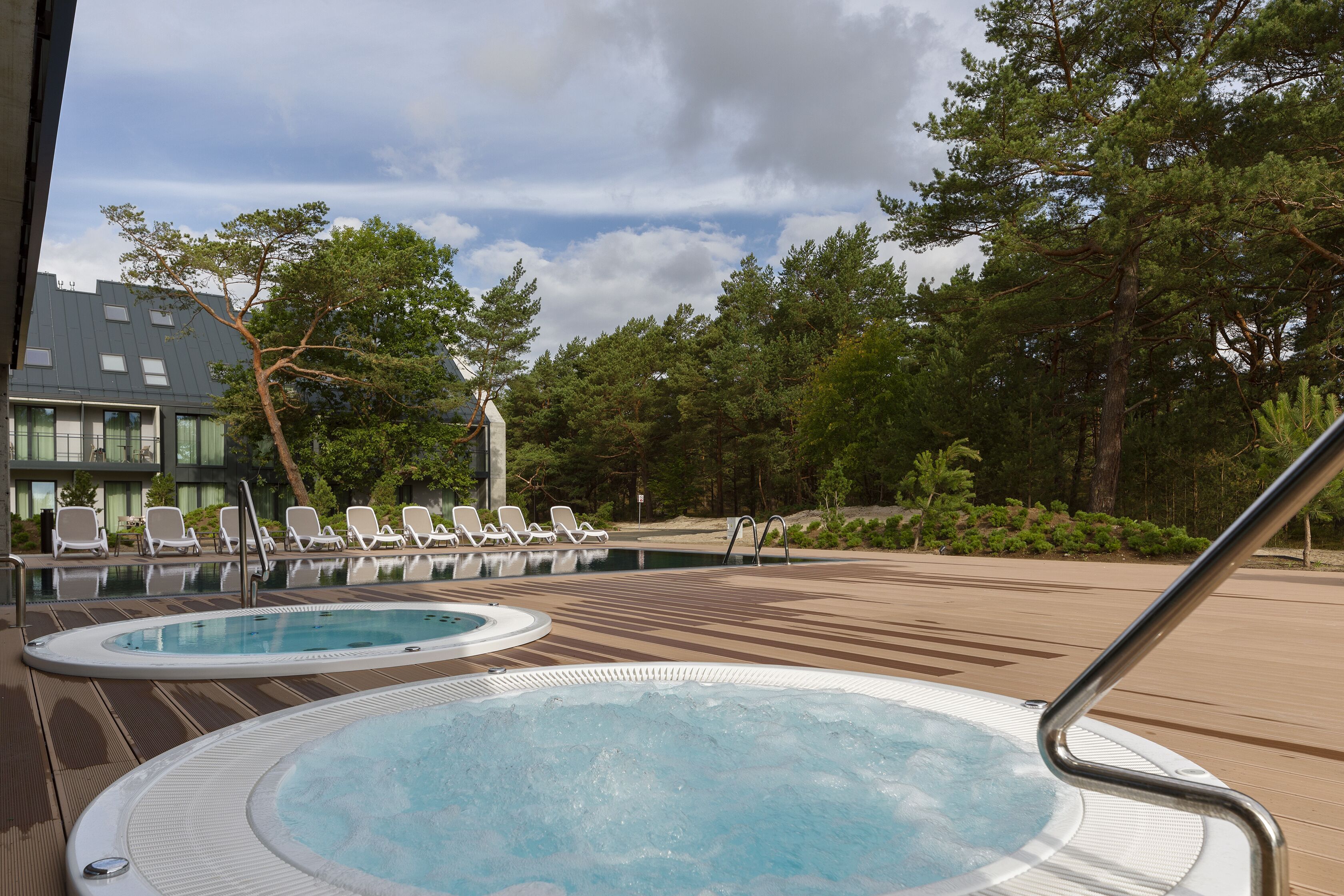 A pool area with hot tubs and lounge chairs, surrounded by trees and a building with large windows.
